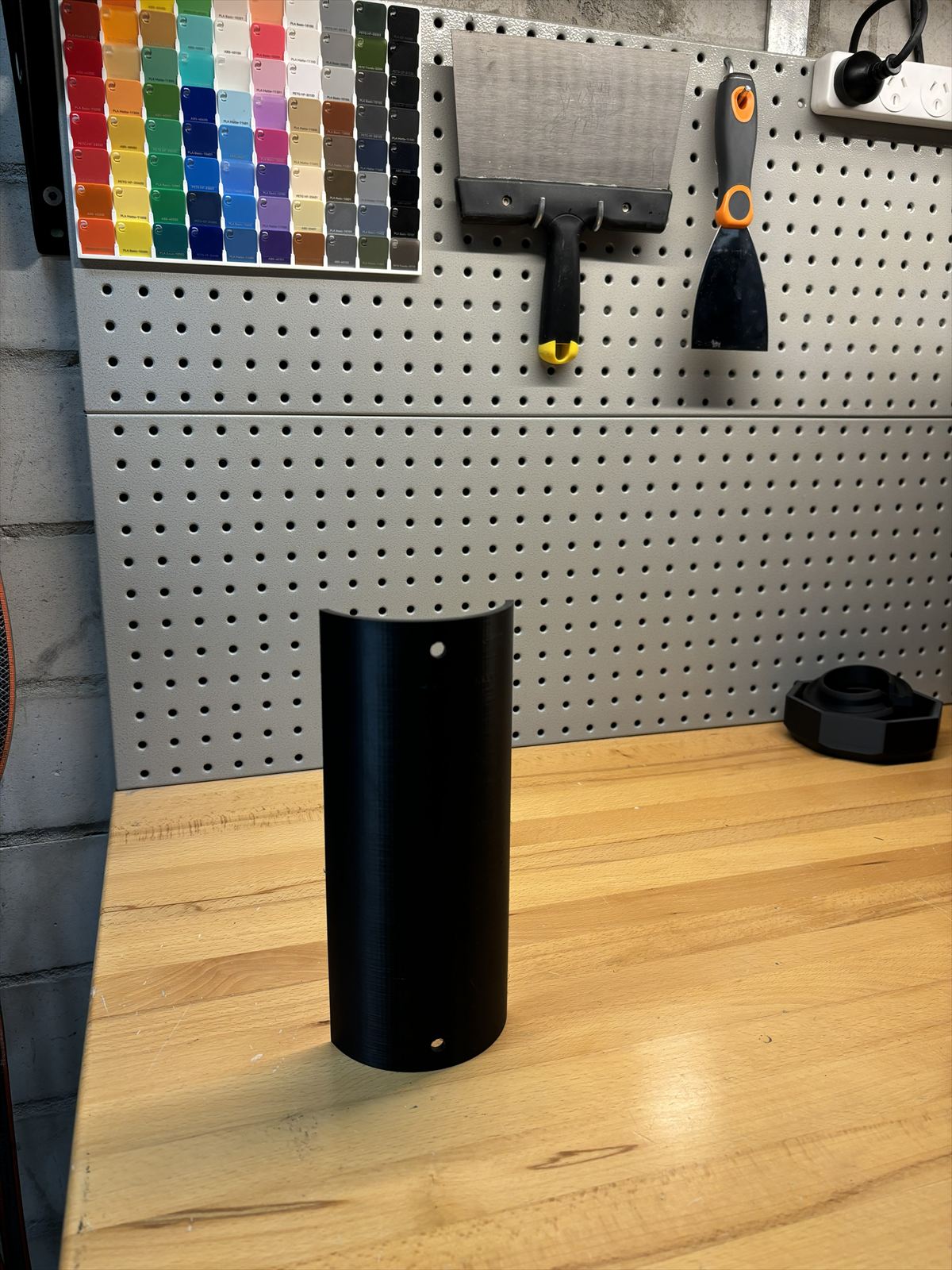 Matte black printed pole cover standing on a timber workbench, colour sample card on the pegboard behind